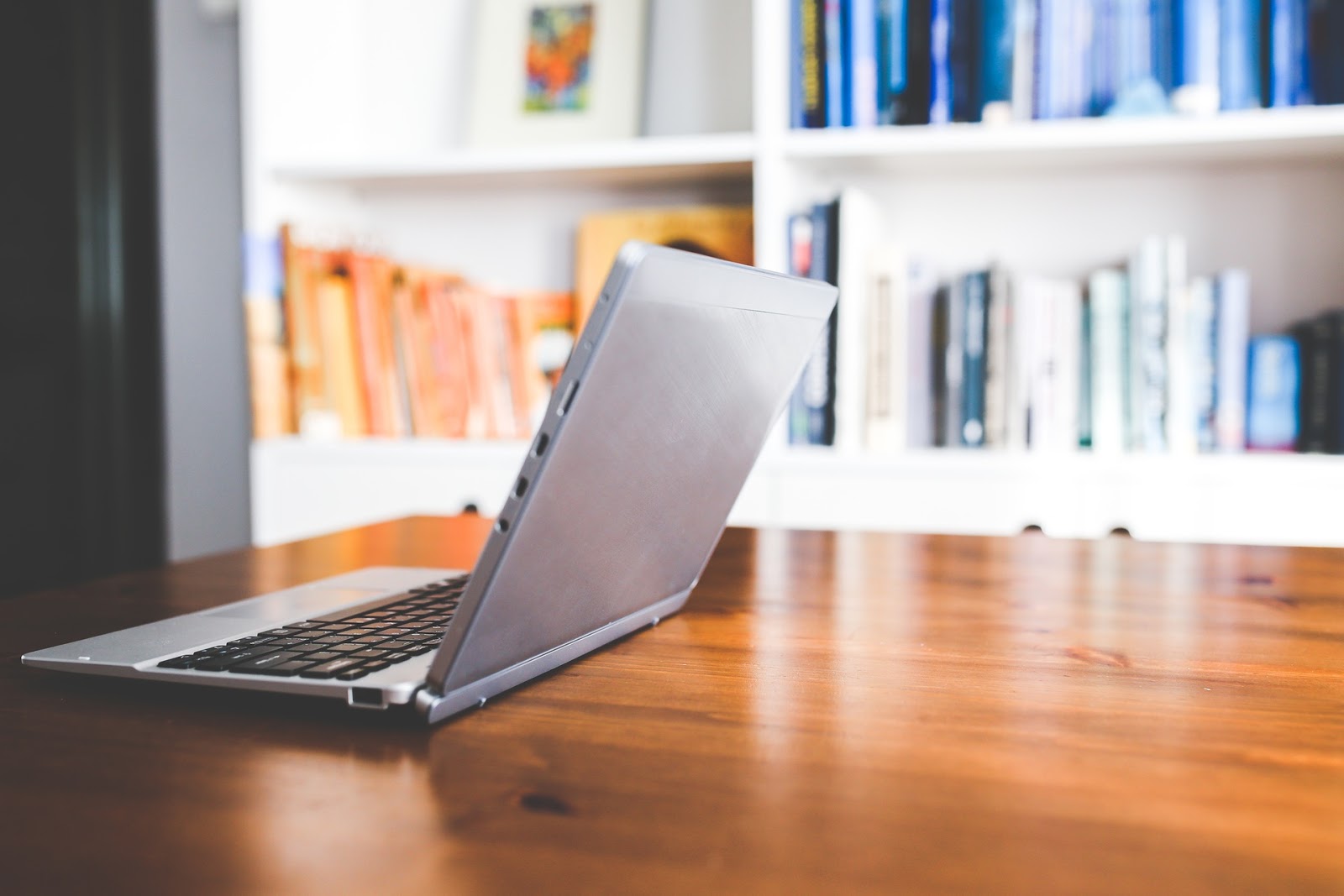 laptop facing away on edge of wooden desk, books on shelf in background in a room filled with natural light