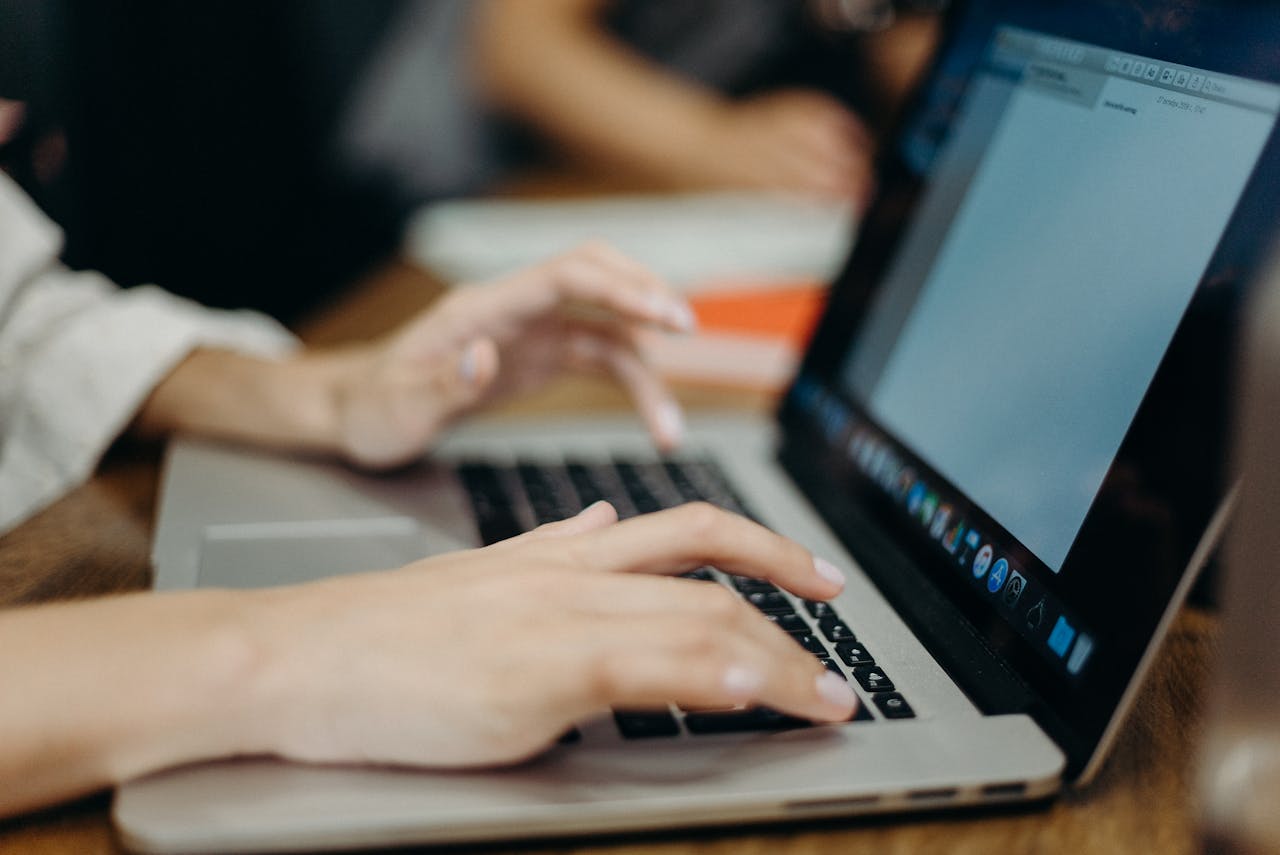 side view of open laptop with hands actively typing, other person working in background